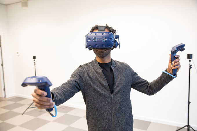 A man wearing a black shirt and gray jacket wears a blue VR headset that covers most of his face over his head. His arms are out and each hand holds a blue controller. He stands in a room with white walls and a checkered floor, and two tripods stand in the background.