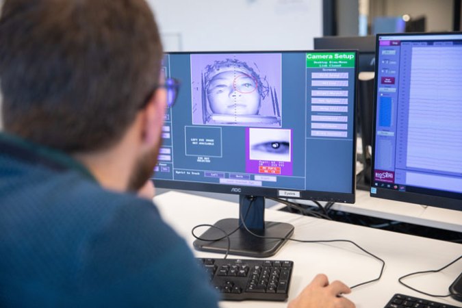 The back of Freek van Ede's head is in the foreground of the picture, blurred and out of focus. The focus of the photograph is on the computer screen he is looking at, which displays a black and white image of the face of the participant in the electroencephalogram (EEG) experiment. There is a red circle around the participant's left eye, and below the face image, there is a magnified image of the eye.